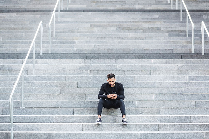 A young adult man sitting outside on a set of steps looking at his phone, reading about the difference between total testosterone and free testosterone